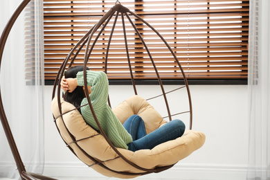 Photo of Young woman relaxing in hanging chair near window at home