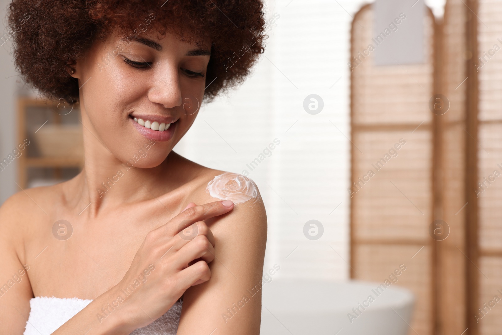 Photo of Beautiful young woman applying body cream onto shoulder in bathroom, space for text