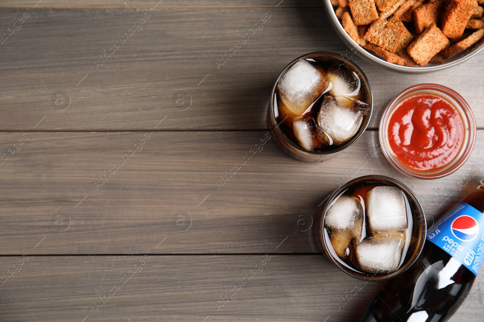 Photo of MYKOLAIV, UKRAINE - FEBRUARY 15, 2021: Glasses and bottle of Pepsi with snack on wooden table, flat lay. Space for text