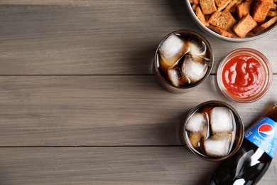 Photo of MYKOLAIV, UKRAINE - FEBRUARY 15, 2021: Glasses and bottle of Pepsi with snack on wooden table, flat lay. Space for text