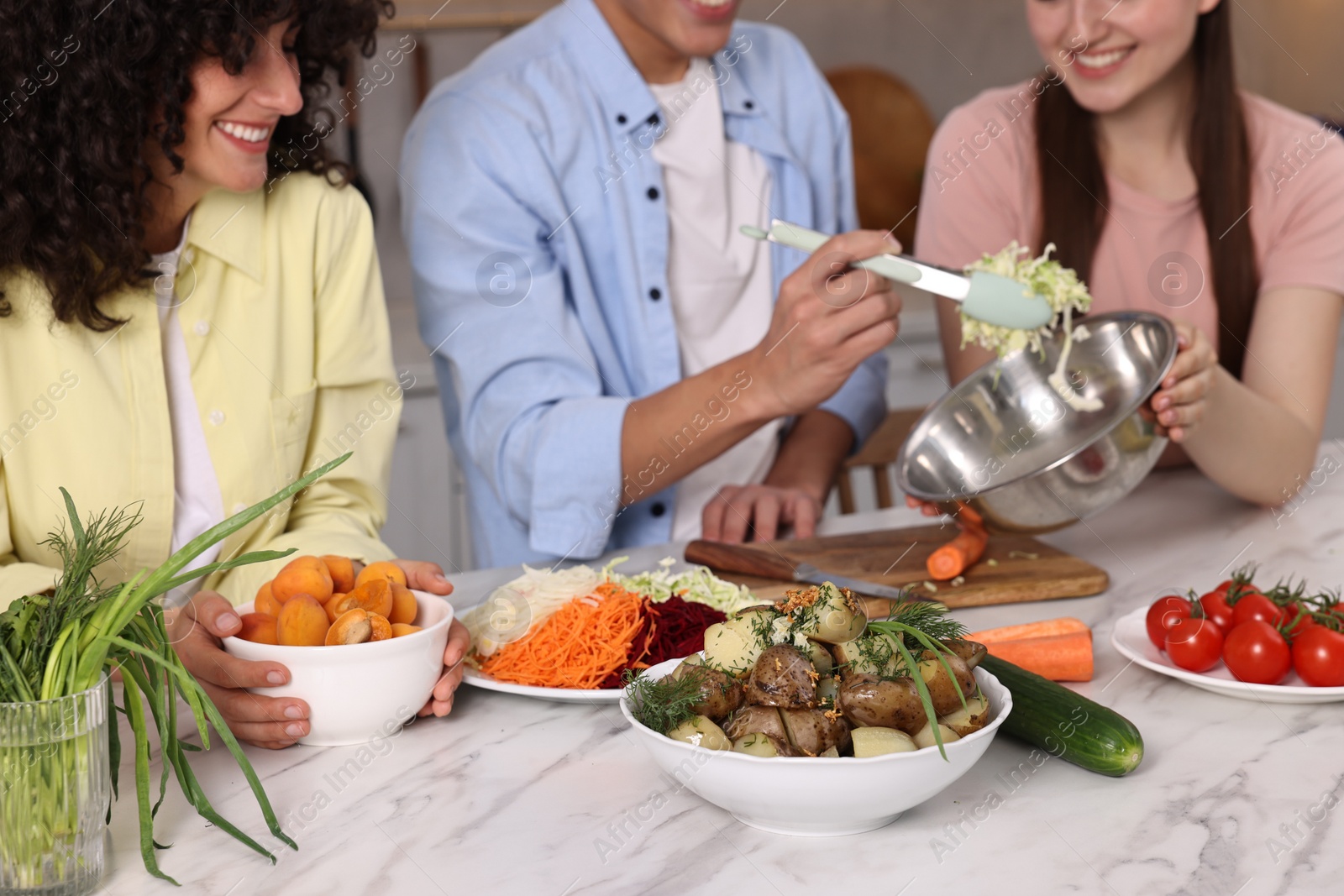 Photo of Friends cooking healthy vegetarian meal at white marble table in kitchen