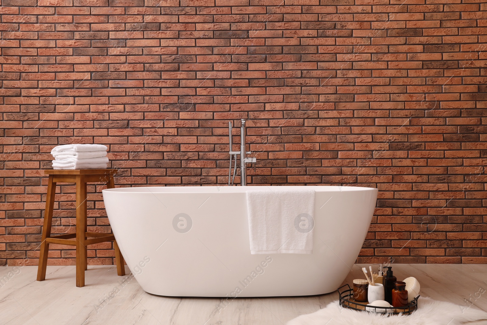 Photo of Modern ceramic bathtub and tray with toiletries near brick wall indoors