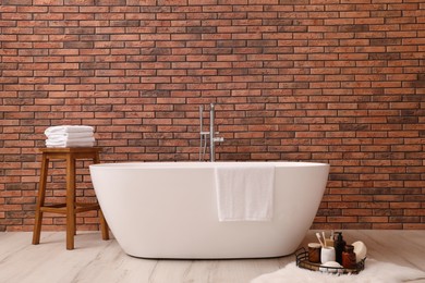 Photo of Modern ceramic bathtub and tray with toiletries near brick wall indoors