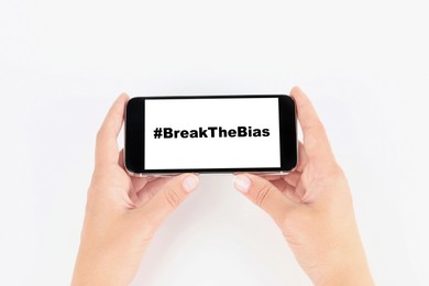 Woman holding smartphone with hashtag BreakTheBias on screen against white background, closeup. Campaign theme for International Women's Day