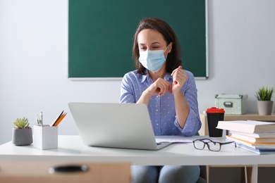 Teacher with protective mask conducting online lesson in classroom during COVID-19 quarantine