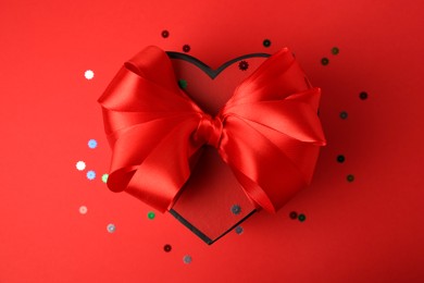 Beautiful heart shaped gift box with bow and confetti on red background, top view