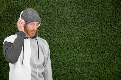 Young man listening to music with headphones near grass wall. Space for text