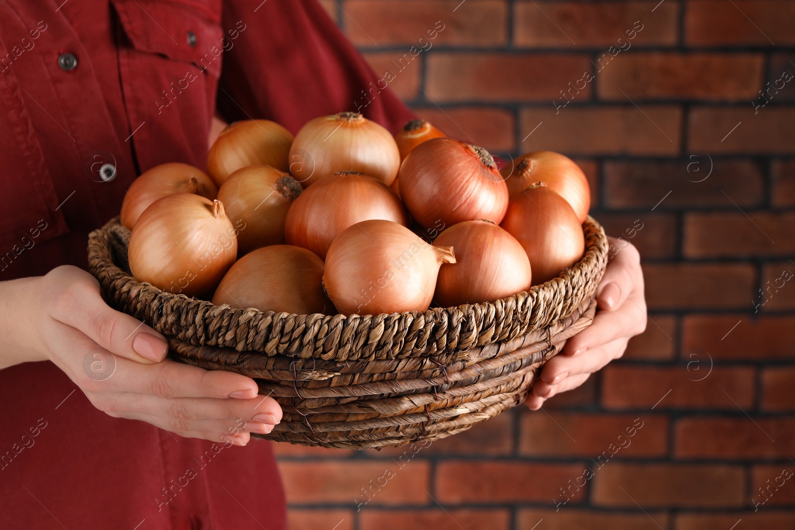 Photo of Woman holding wicker basket with ripe onions against red brick wall, closeup