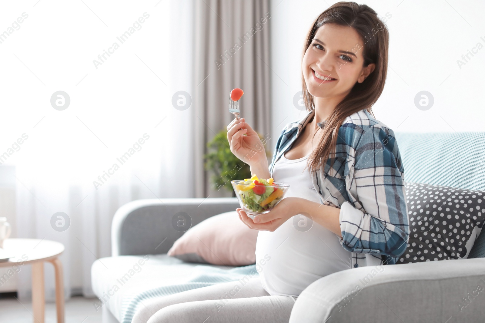 Photo of Young pregnant woman eating vegetable salad on sofa at home