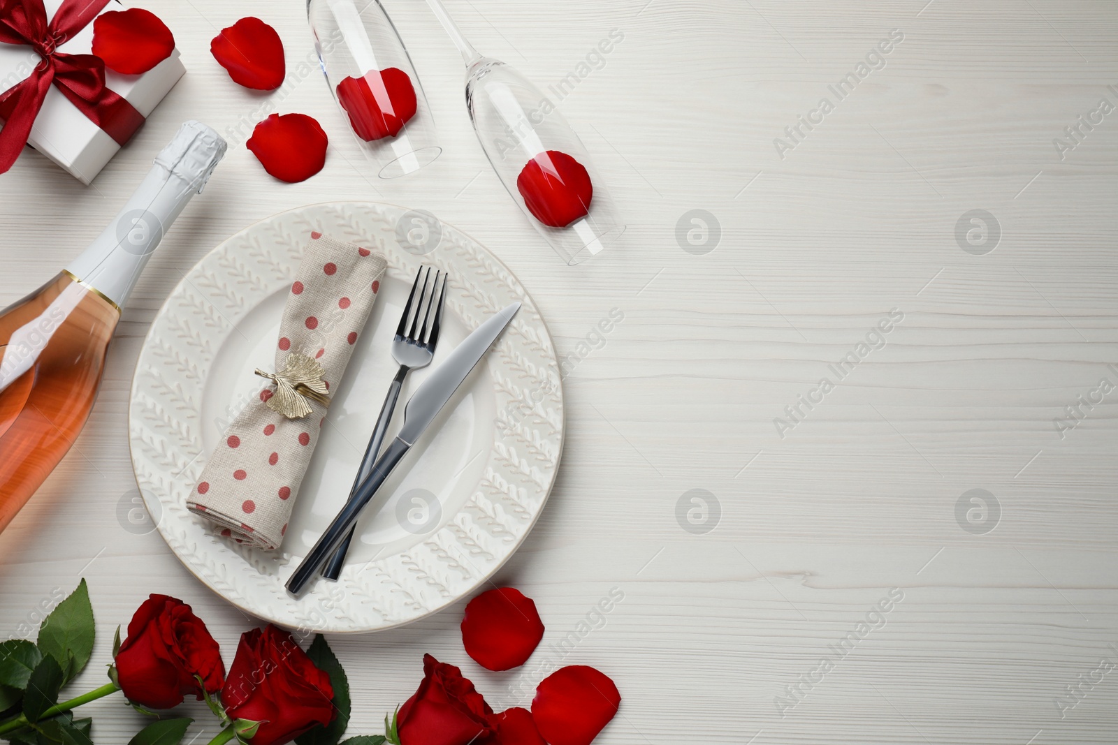 Photo of Beautiful table setting with gift box and rose petals on white wooden table for romantic dinner, flat lay. Space for text