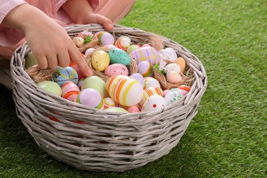 Photo of Child with wicker basket full of Easter eggs on green grass, closeup