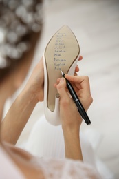 Photo of Young bride writing her single friends names on shoe indoors, closeup. Wedding superstition