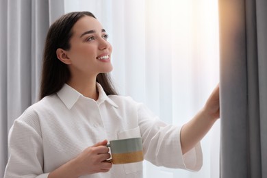 Photo of Happy woman with cup of drink near window at home, space for text. Lazy morning