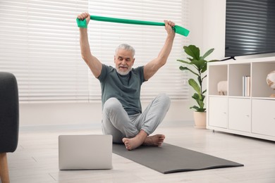 Photo of Senior man doing exercise with fitness elastic band near laptop on mat at home