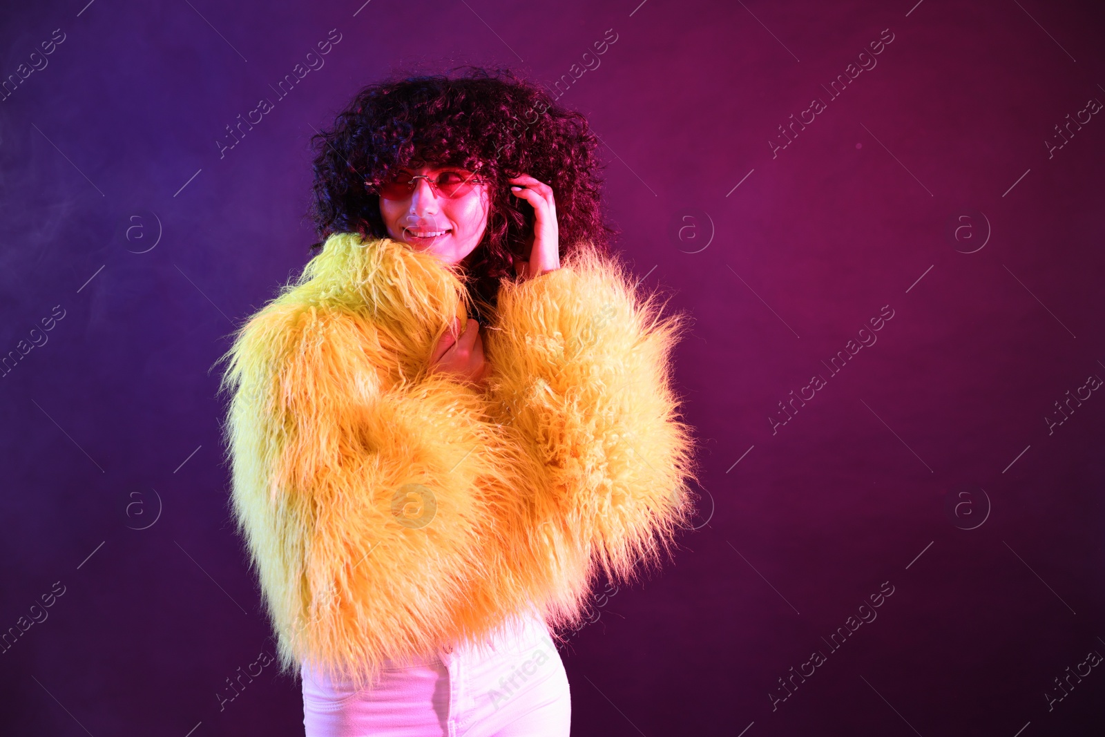 Photo of Beautiful young woman in yellow fur coat and sunglasses on color background in neon lights. Space for text