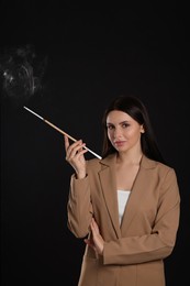 Woman using long cigarette holder for smoking on black background, space for text