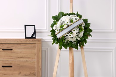 Wreath of flowers and photo frame with black ribbon on commode in room. Funeral attributes