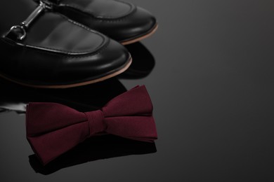 Photo of Stylish burgundy bow tie and shoes on black mirror surface, space for text