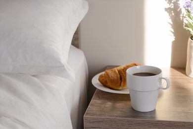 Photo of Cup of morning coffee and croissant on wooden night stand near bed indoors