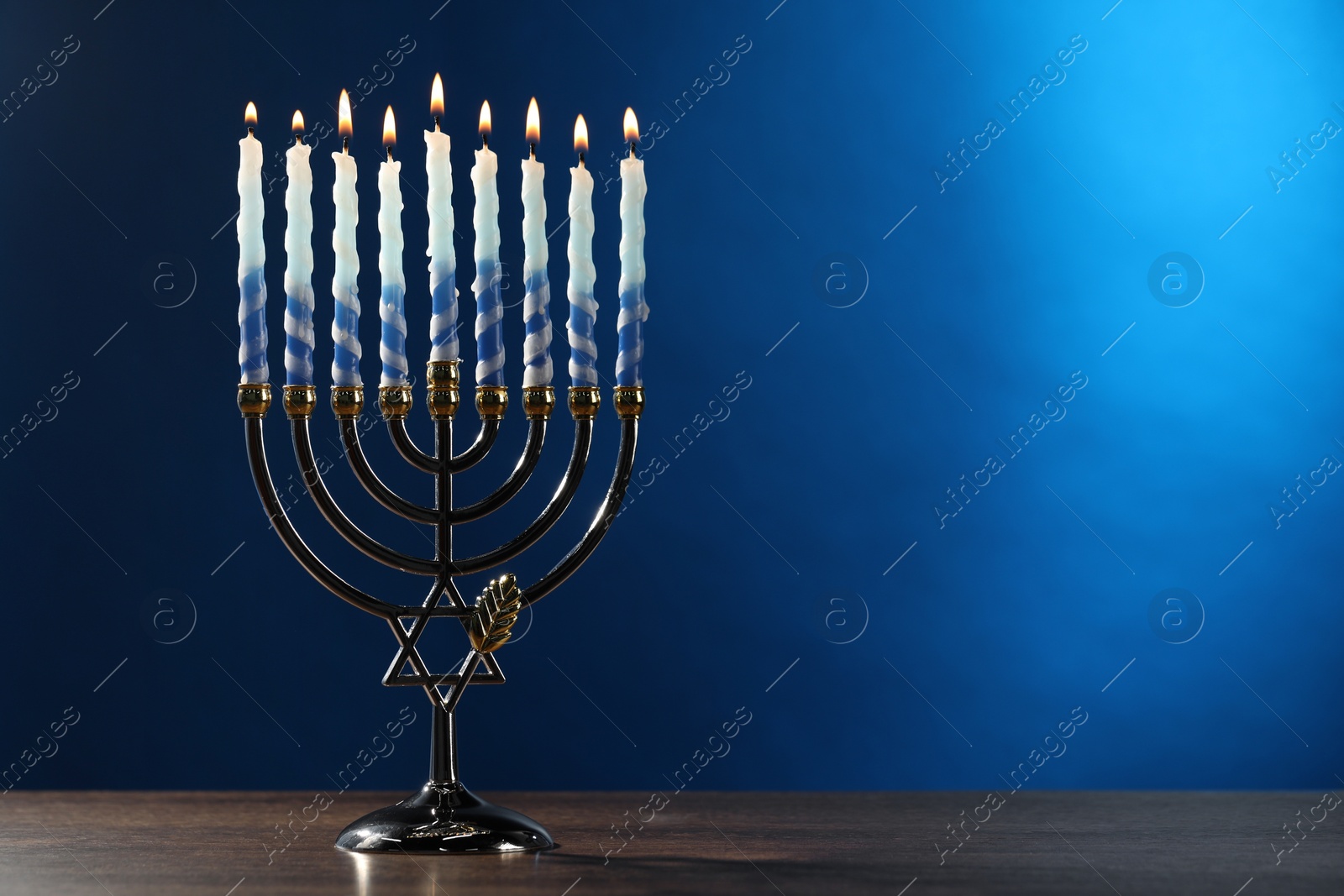 Photo of Hanukkah celebration. Menorah with burning candles on wooden table against blue background, space for text