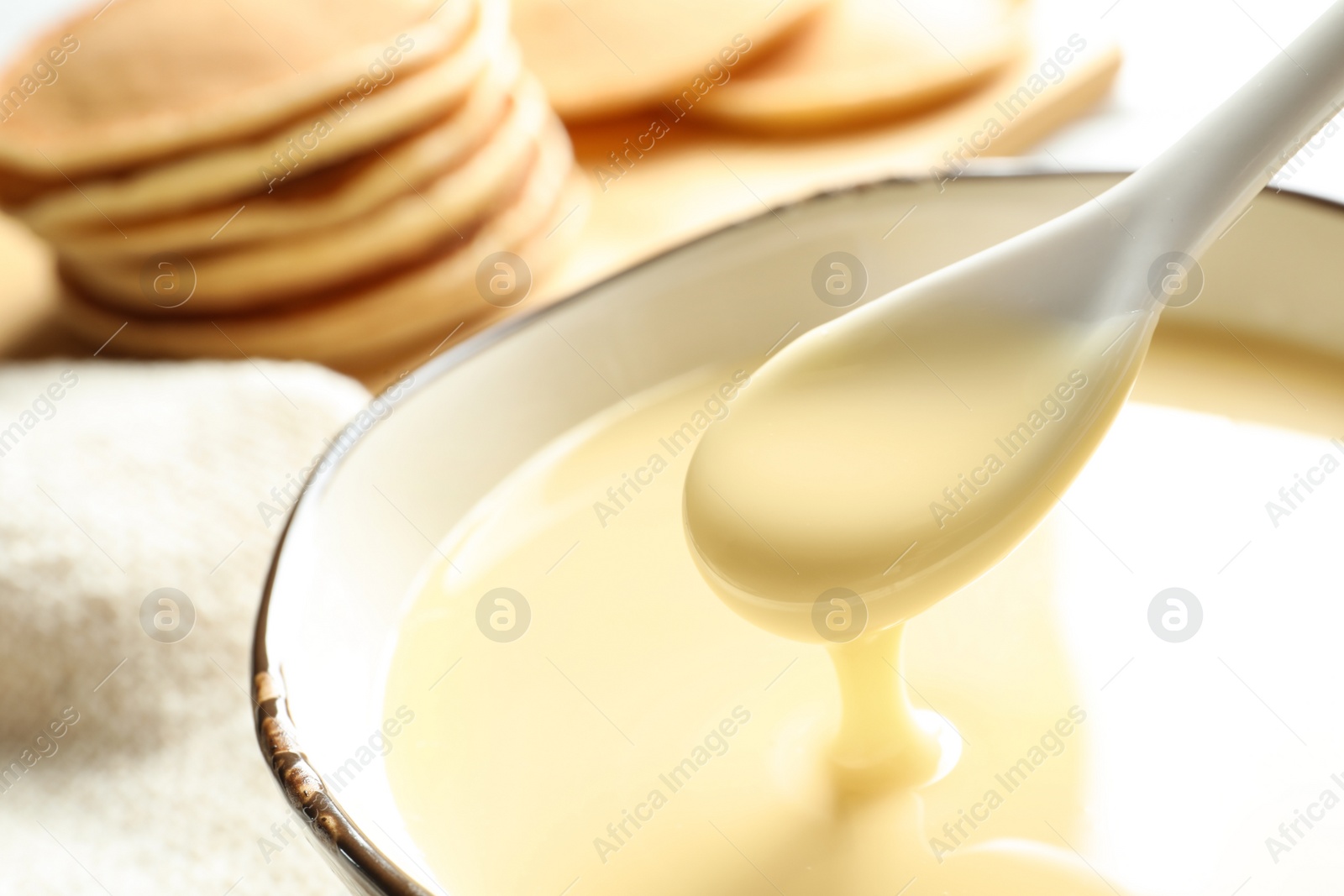 Photo of Spoon of pouring condensed milk over bowl on table, closeup with space for text. Dairy products