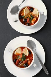 Meat solyanka soup with sausages, olives, vegetables and spoons on dark grey textured table, flat lay