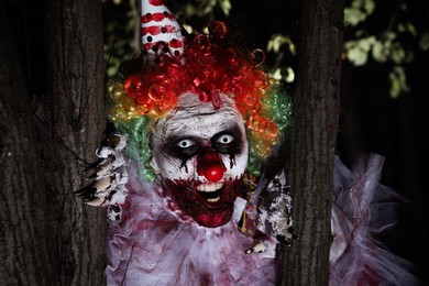 Photo of Terrifying clown hiding behind trees outdoors at night. Halloween party costume