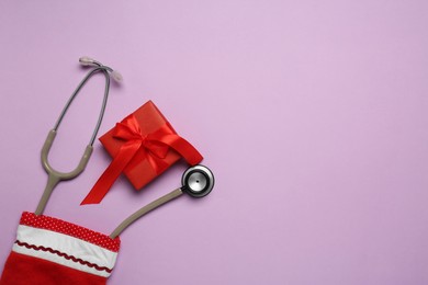 Photo of Greeting card for doctor with stethoscope and Christmas gift on purple background, flat lay. Space for text