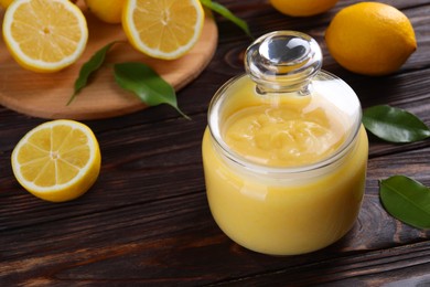 Photo of Delicious lemon curd in glass jar, fresh citrus fruits and green leaves on wooden table