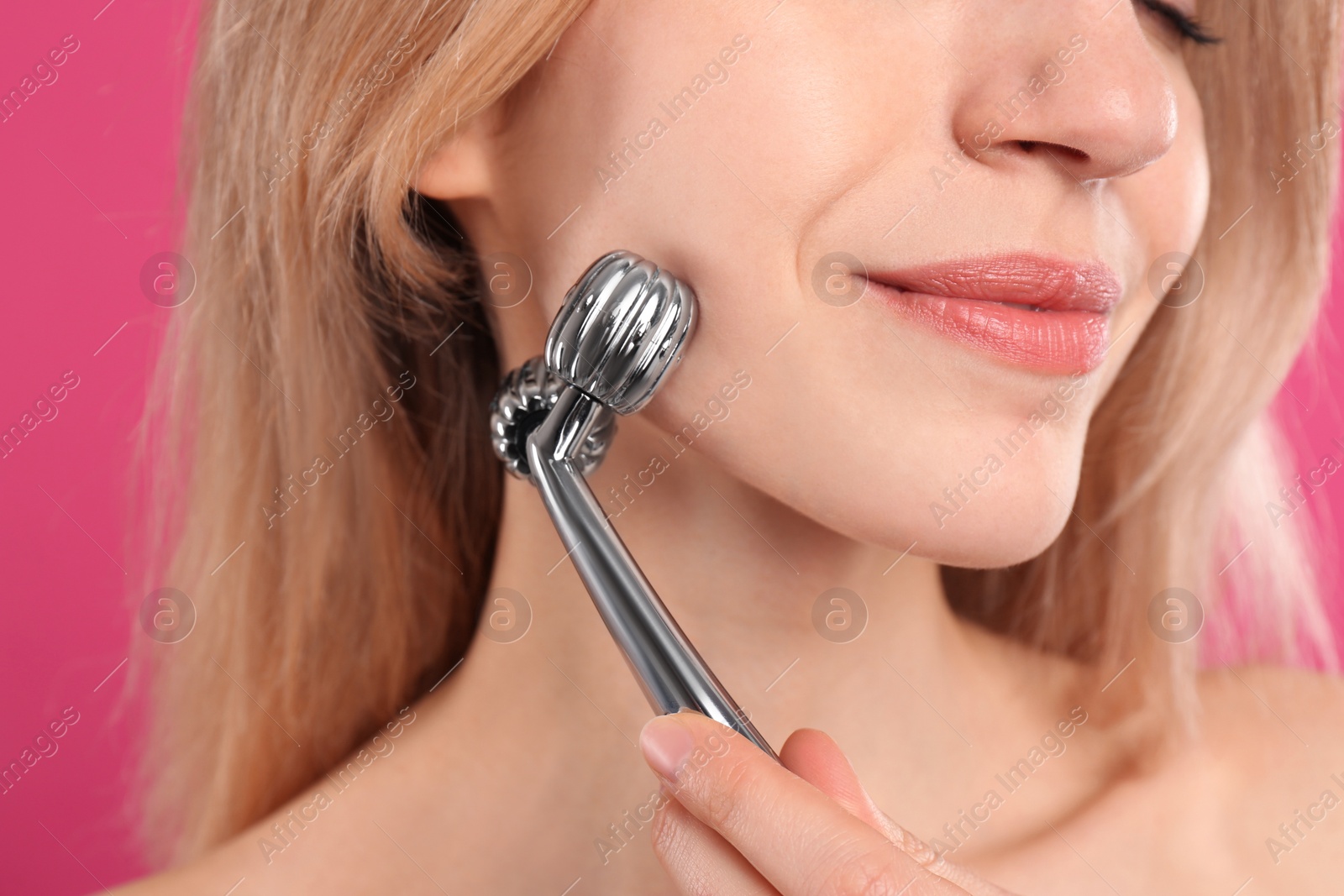 Photo of Young woman using metal face roller on pink background, closeup