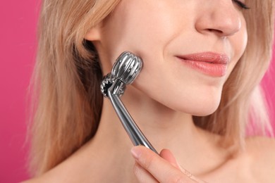Young woman using metal face roller on pink background, closeup