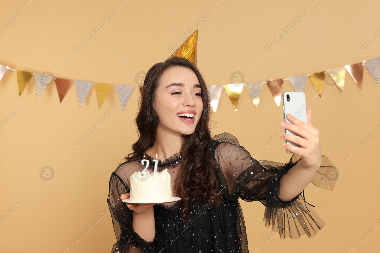 Photo of Coming of age party - 21st birthday. Smiling woman holding delicious cake with number shaped candles and taking selfie against beige background