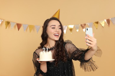 Coming of age party - 21st birthday. Smiling woman holding delicious cake with number shaped candles and taking selfie against beige background