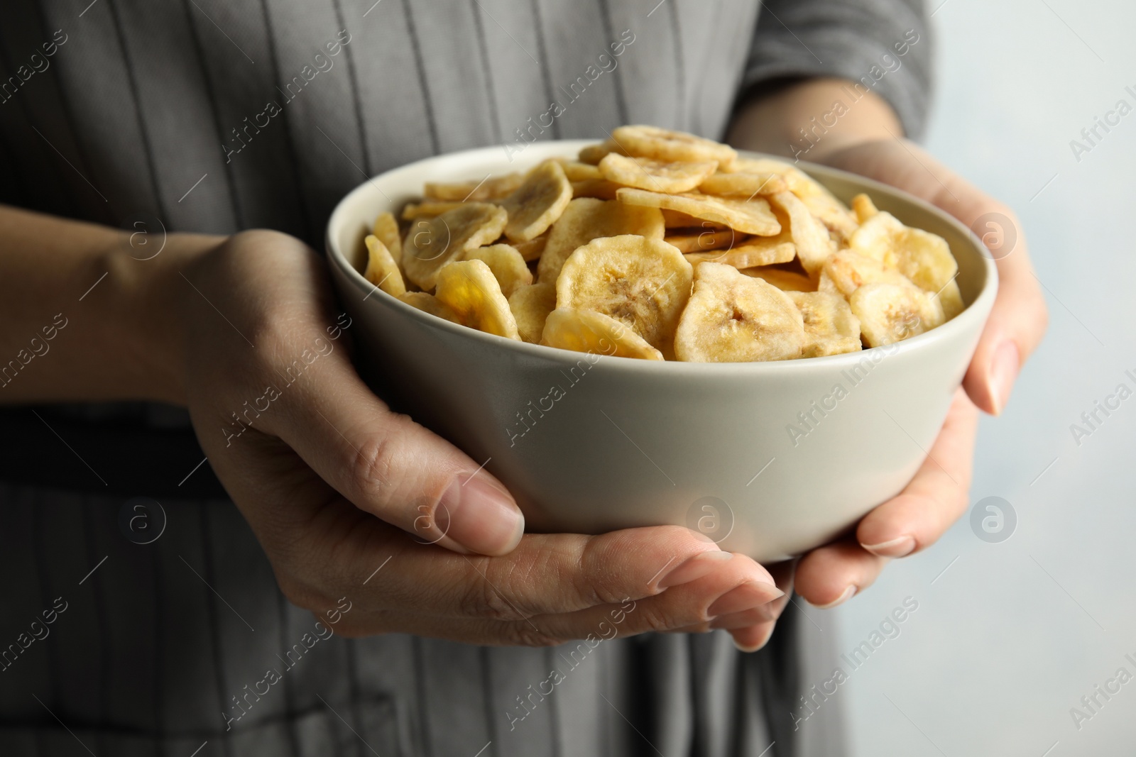 Photo of Woman holding bowl with sweet banana slices on light background, closeup. Dried fruit as healthy snack