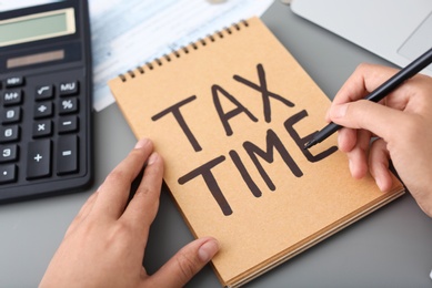 Photo of Woman writing TAX TIME on notebook at table, closeup