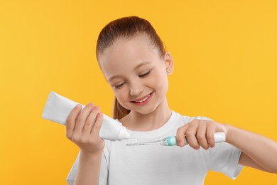 Photo of Happy girl squeezing toothpaste from tube onto electric toothbrush on yellow background