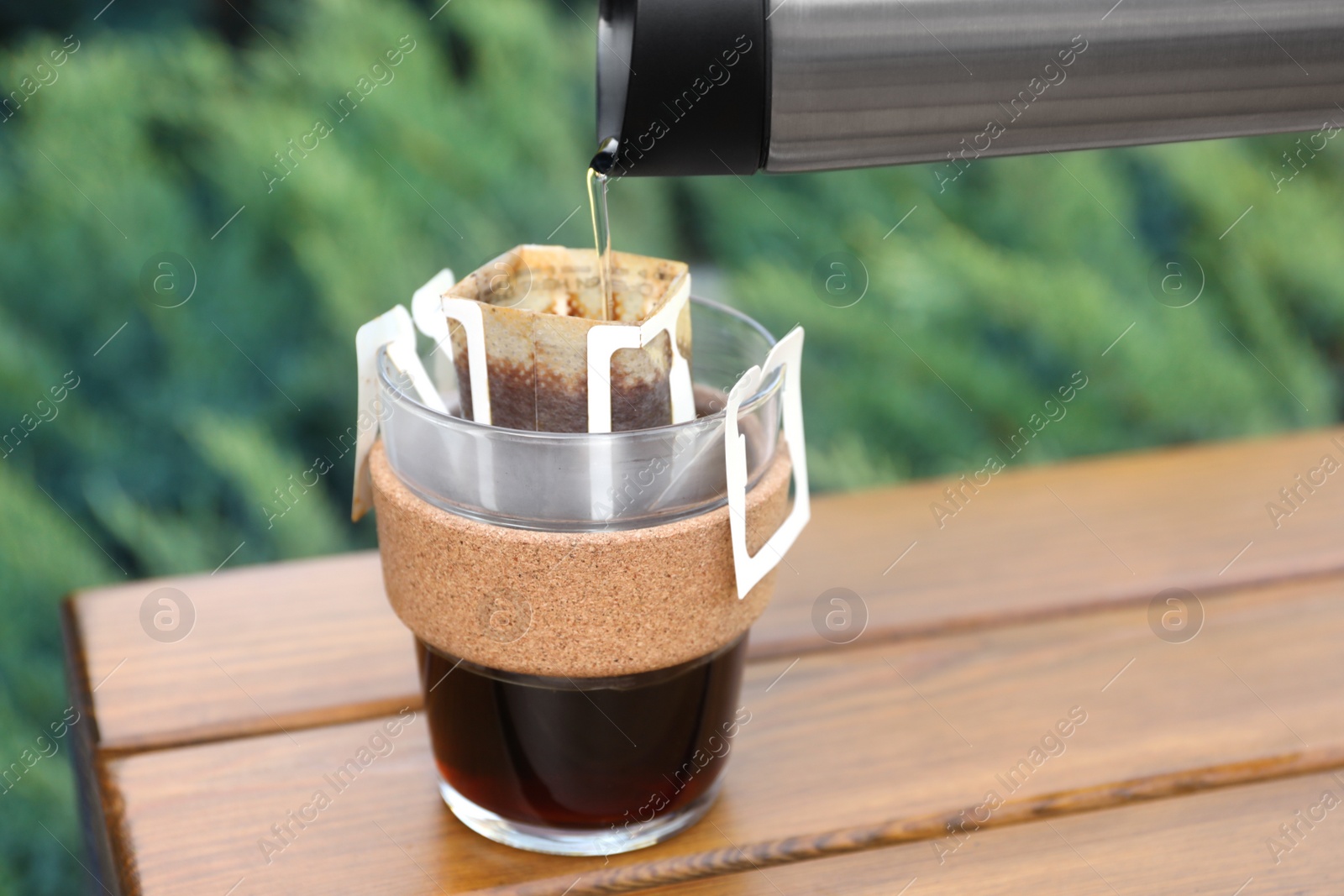 Photo of Pouring hot water into glass with drip coffee bag from thermos on wooden table, closeup