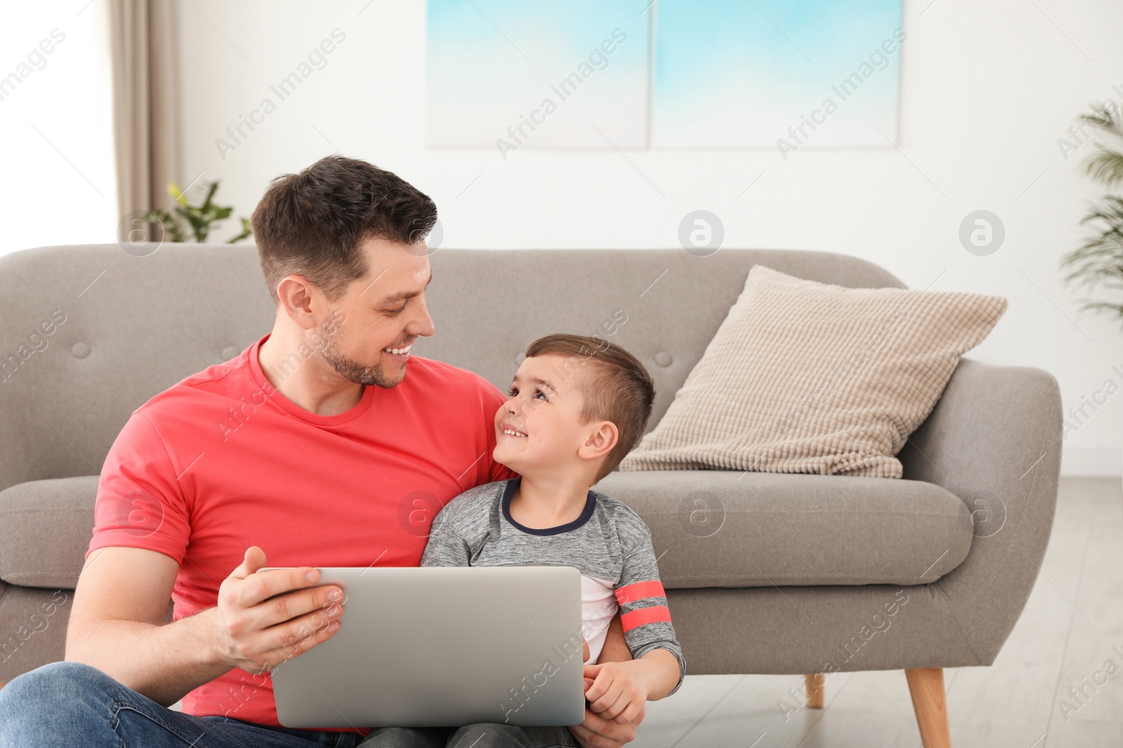 Photo of Boy and his father with laptop sitting near the sofa on floor. Family time