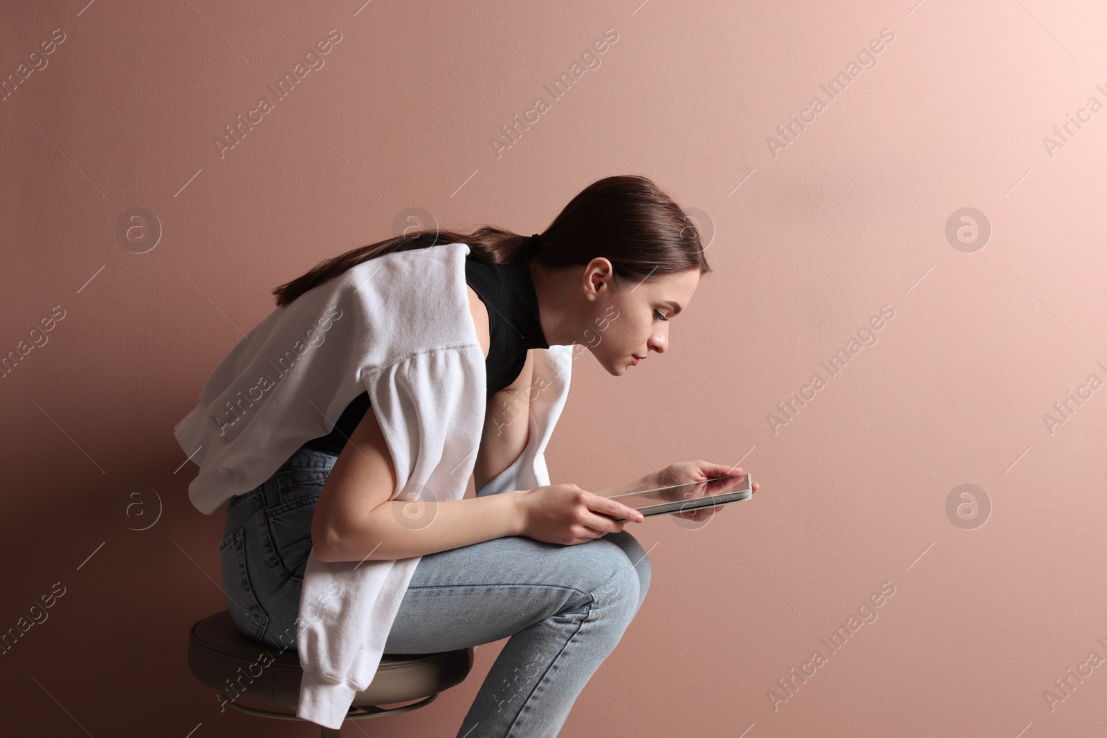 Photo of Young woman with bad posture using tablet while sitting on stool against pale pink background