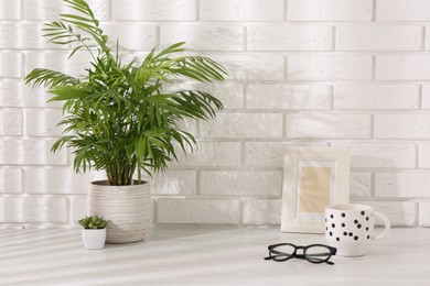 Photo of Stylish office workplace. Frame, plants, glasses and cup on white table near brick wall