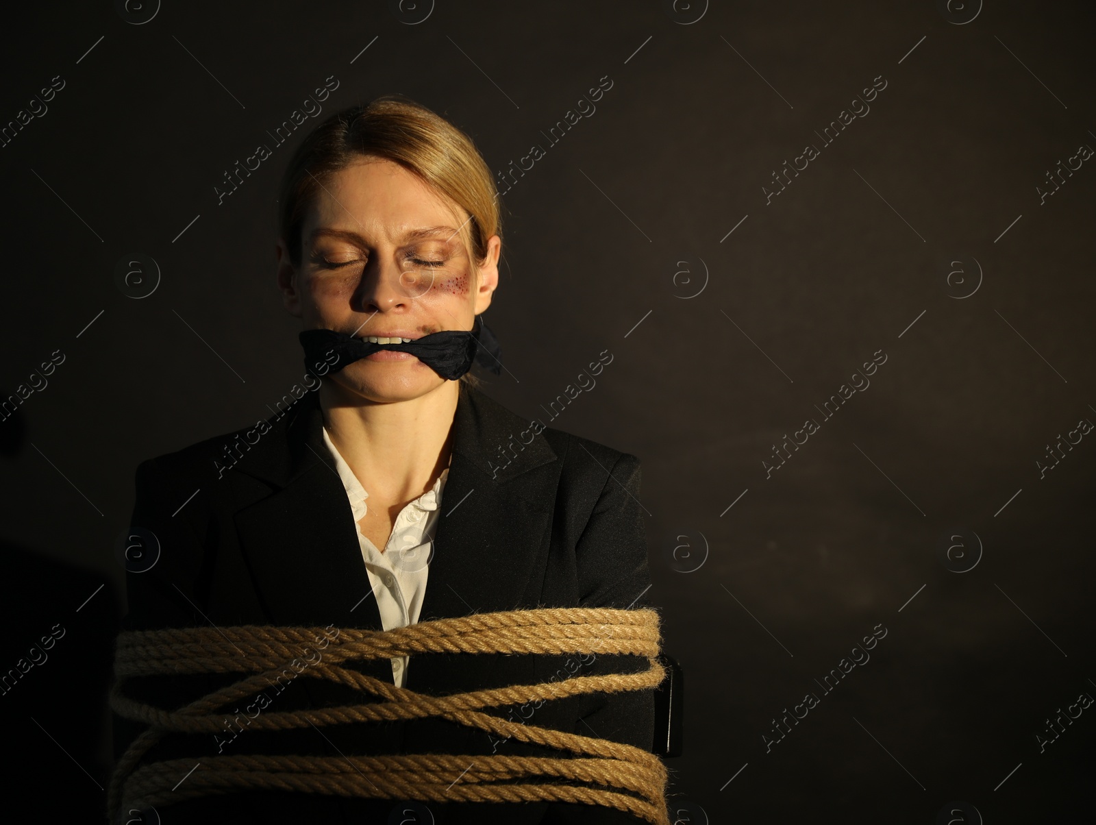 Photo of Woman with bruises tied up and taken hostage on dark background. Space for text