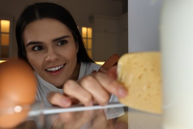 Young woman taking cheese out of refrigerator in kitchen at night, view from inside
