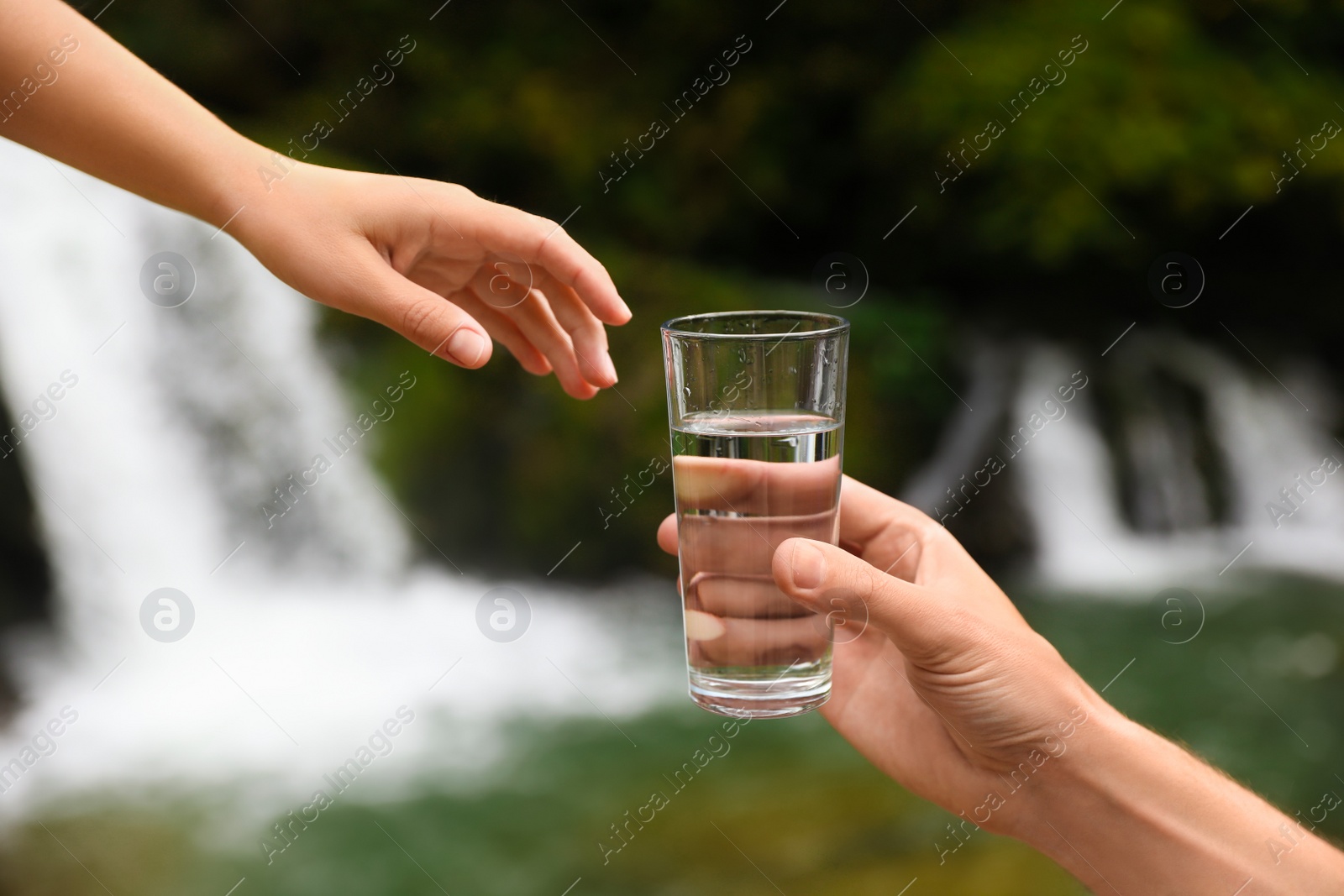 Photo of Man giving woman glass of fresh water near waterfall, closeup