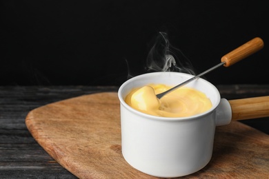 Pot of tasty cheese fondue and fork with bread on black wooden table