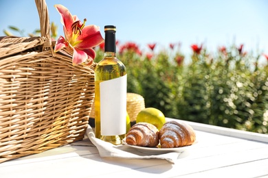 Photo of Composition with picnic basket and bottle of wine on white wooden table in lily field. Space for text