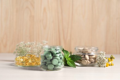 Photo of Jars with different pills, flowers and mint on white wooden table, space for text. Dietary supplements