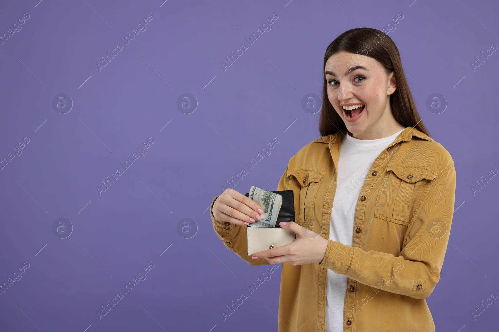 Photo of Excited woman putting dollar banknotes into wallet on purple background, space for text