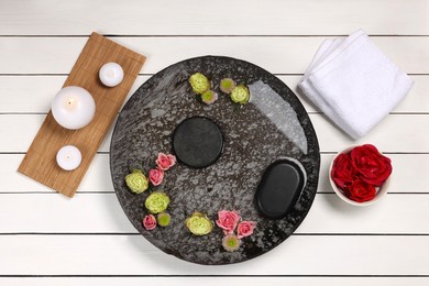 Photo of Plate with water, flowers, stones and burning candles on white wooden floor, flat lay. Pedicure procedure