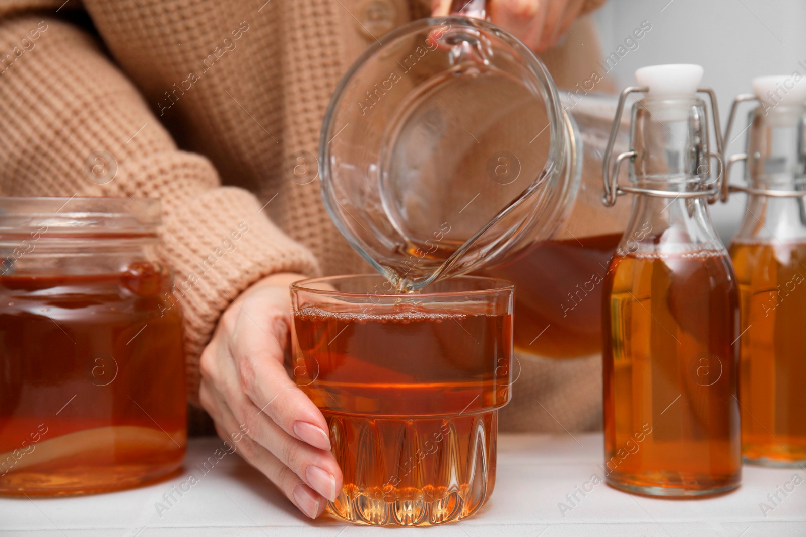 Photo of Woman pouring fermented kombucha from jug into glass at white table, closeup