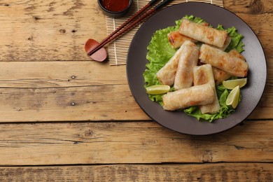 Photo of Plate with tasty fried spring rolls, lettuce, lime, sauce and chopsticks on wooden table, top view. Space for text